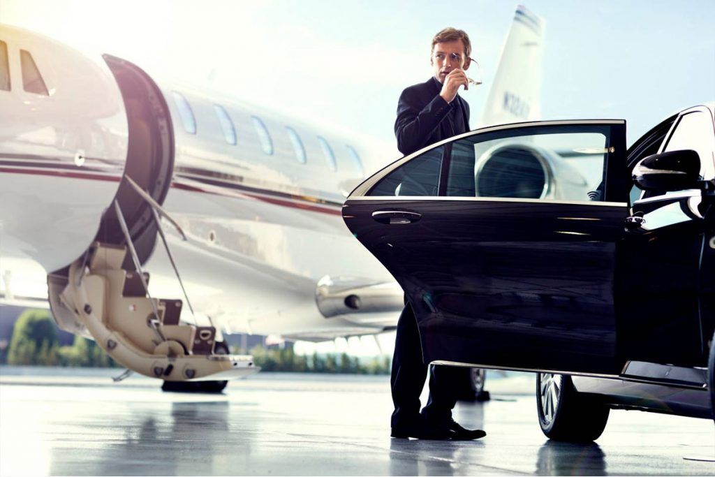 A businessman stepping out of a chauffeured luxury car next to a private jet on an airport tarmac.