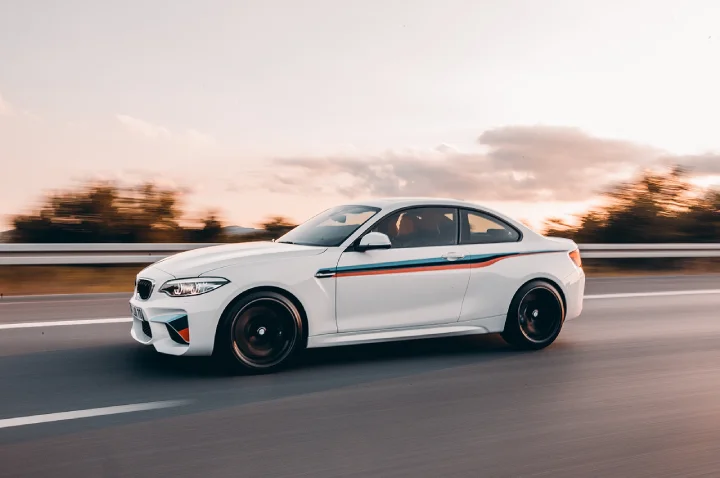 White performance coupe with racing stripes cruising on a highway, featured in our luxury car rental fleet.